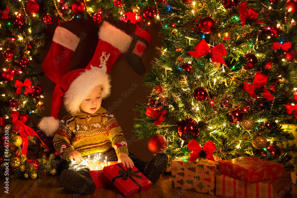 Christmas Kid opening Gift Box with Xmas Lights Garland. Cute Baby in Santa Hat sitting next to Decorated Fir Tree in Dark Room Interior with Shining Ornaments