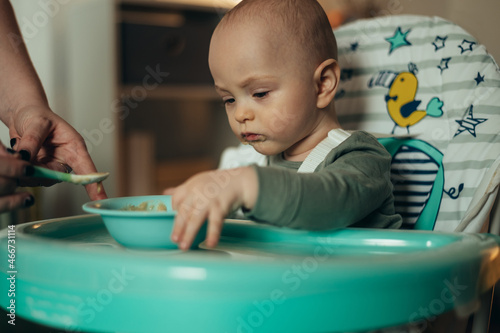 Baby eating with a face stained in food while mother feeds him with a spoon