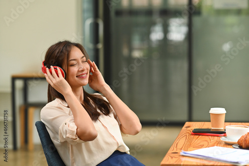 Photo of a young beautiful woman wearing headphones relaxing at the comfortable wooden table surrounded by breakfast and office equipment.