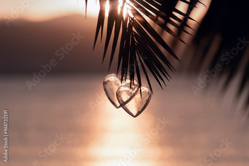 Fototapeta Naklejka Na Ścianę i Meble -  Two decorative hearts hanging on palm tree branch against background of romantic sunset over sea beach illuminated by sunlight. Couple in love. Christmas holiday, Valentine's Day concept