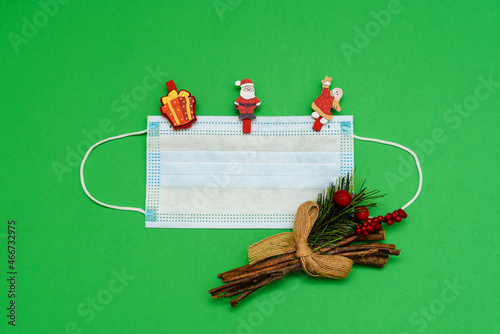 Medical mask on green background with spruce branch and pins - toys in the form of gift, Santa Claus and angel. Top view with copy space, space for text. Happy New Year 2022 concept.