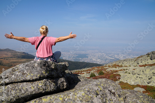 blonde in a pink t-shirt sits on the top of the mountain, arms outstretched to the sides