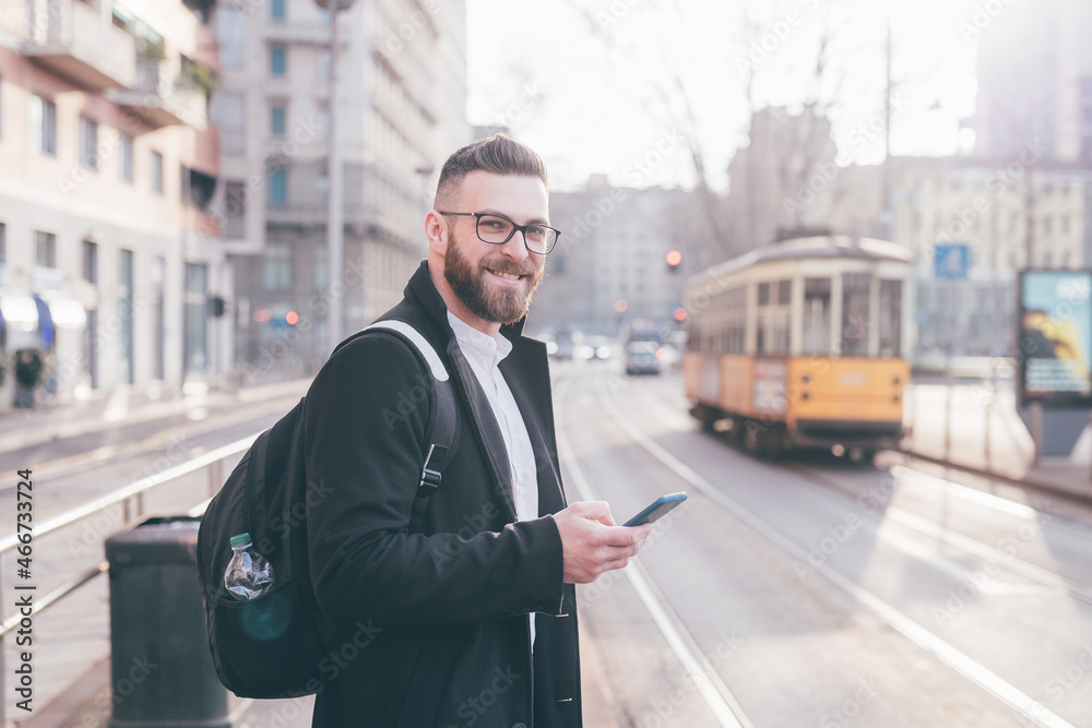 Smiling commuter contemporary bearded businessman outdoors at bus station using smartphone smiling positive Smiling commuter contemporary bearded businessman outdoors at bus station using smartphone smiling positive