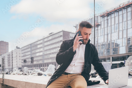 Young caucasian bearded contemporary businessman freelancer remote working outdoor city talking smartphone using computer photo