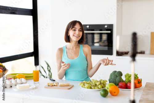 Food blogger concept. Millennial woman recording new video recipe on smartphone  standing in modern kitchen interior