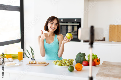Happy woman showing thumb up and holding apple, recording new video for sport and food blog, kitchen interior