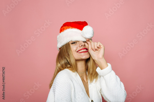 A funny woman covers one eye with a Santa hat and laughs. Flirty lady on a pink background celebrates Christmas and New Year