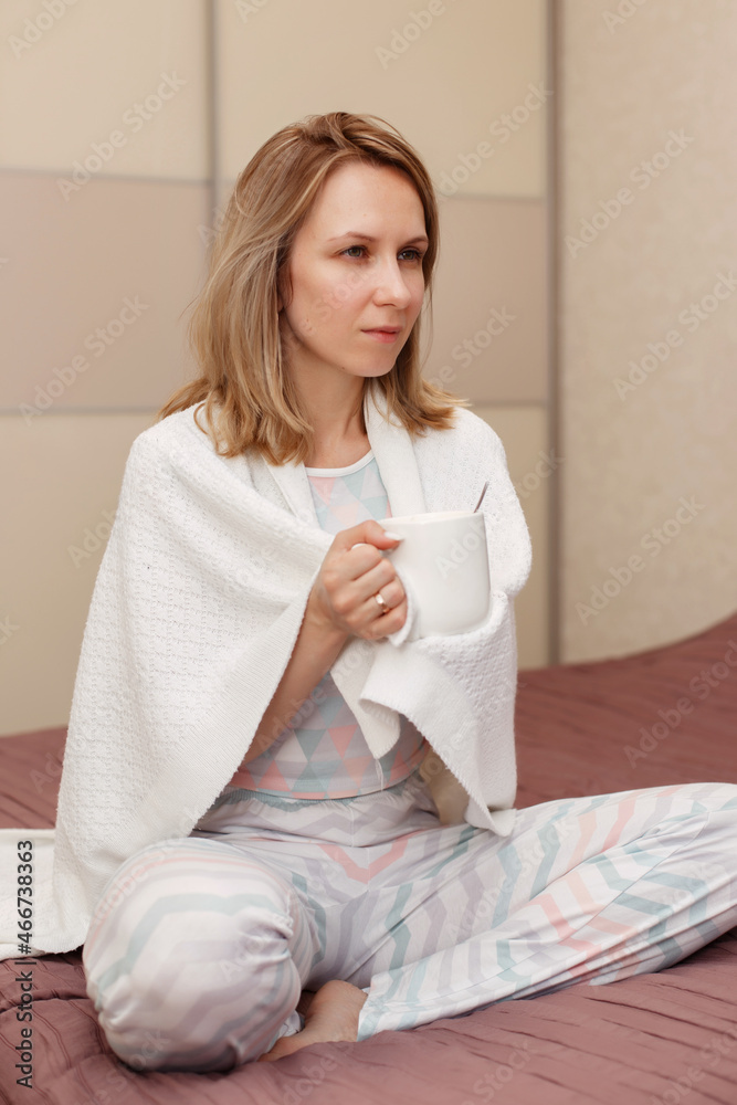 The girl is holding a white cup of tea or coffee with marshmallows. A model in a white sweater and a cozy plaid is sitting, resting at home on the sofa. The girl is holding a white cup of tea or coffee with marshmallows. A model in a white sweater and a cozy plaid is sitting, resting at home on the sofa.