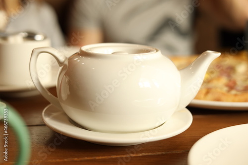 A white teapot with healthy herbal tea on the table in a cafe, served to guests in a restaurant or cafe. Pizza in the background. Relax and unwind