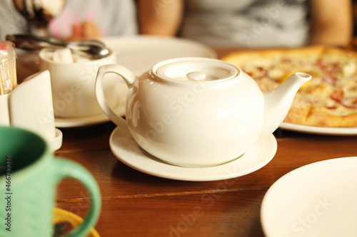 A white teapot with healthy herbal tea on the table in a cafe, served to guests in a restaurant or cafe. Pizza in the background. Relax and unwind