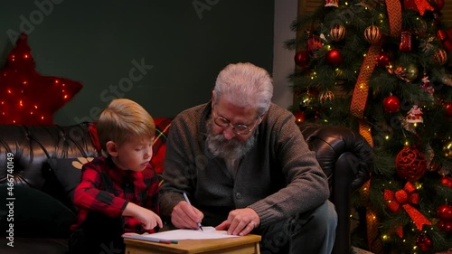 Grandfather teaches his grandson to draw with felttip pens. An elegant gray haired man with a little boy is sitting on a sofa in a decorated room near a glowing Christmas tree. Close up. Slow motion. photo
