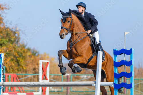 Fotografiet Young sportsman riding the sorrel horse jumping over an obstacle on show jumping competition