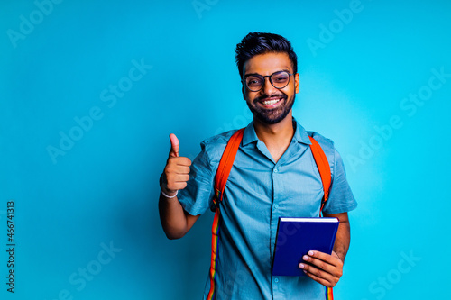 handsome young bearbed indian man with eye glasses in blue cotton t-shirt with orange rainbow backpack i thumbs up n studio background photo