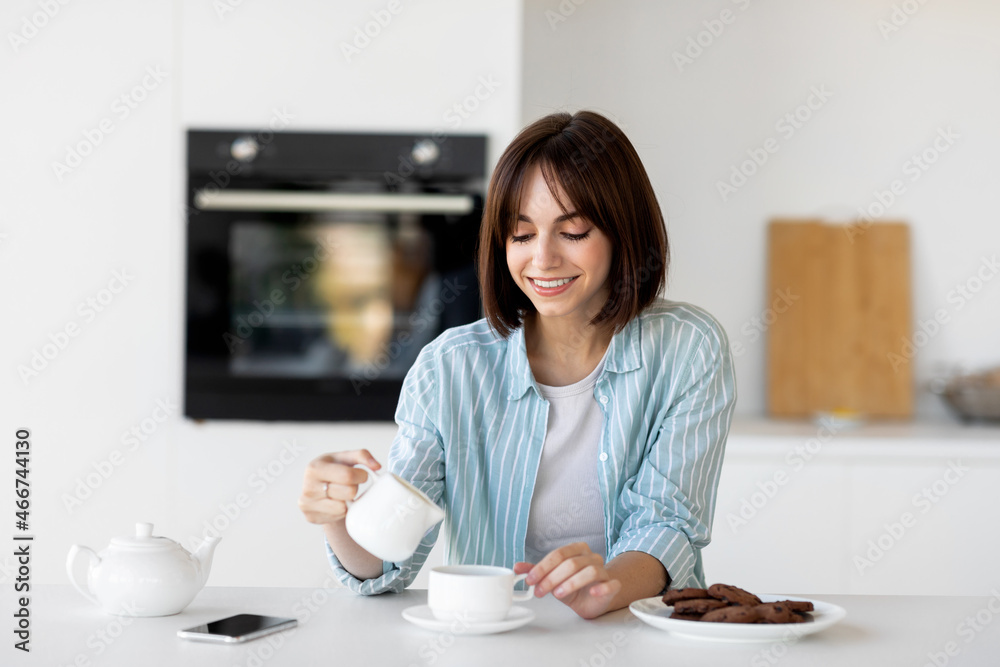 Happy young lady pouring milk in cup of coffee, sitting in kitchen interior, enjoying morning hot beverage Happy young lady pouring milk in cup of coffee, sitting in kitchen interior, enjoying morning hot beverage
