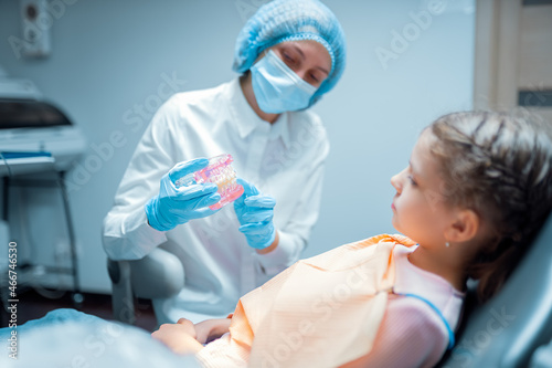 female pediatric dentist orthodontist teaching little girl patient using teeth model with brecets.