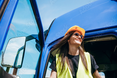 Portrait of beautiful young woman professional truck driver with protective yellow helmet sitting and driving a big truck. Inside of vehicle. People and industrial transportation concept.