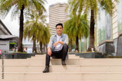 Full length portrait of handsome young man sitting outdoors in city during summer