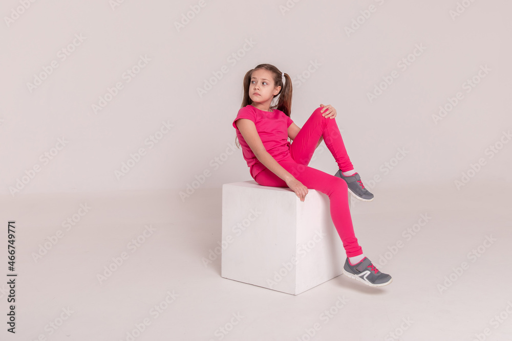 beautiful girl in a sports pink T-shirt and pants on a white isolated background. child sitting on a white cube beautiful girl in a sports pink T-shirt and pants on a white isolated background. child sitting on a white cube