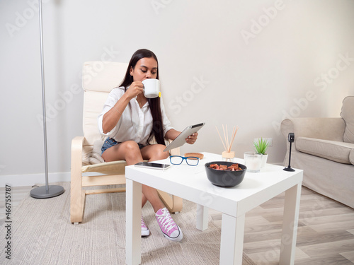 Young woman at home tired from work with a tablet sitting on a chair with a table in front of her in a relaxed atmosphere and drinking tea