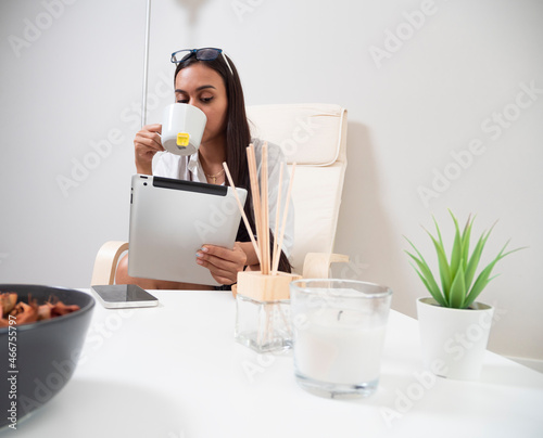 Young woman at home tired from work with a tablet sitting on a chair with a table in front of her in a relaxed atmosphere and drinking tea