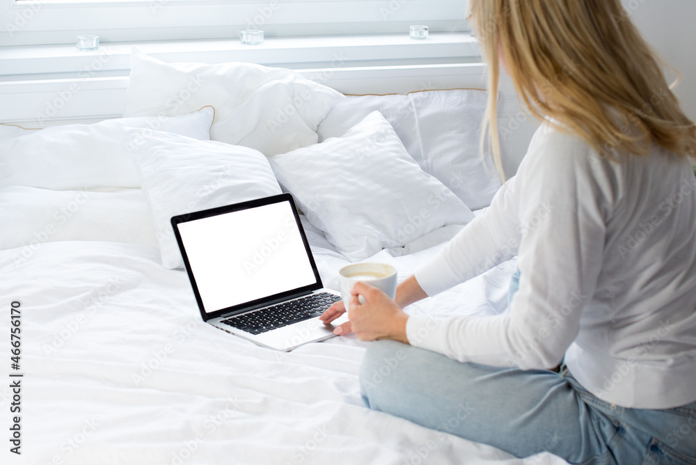Blonde woman is sitting on the white bed and looks at the mockup laptop. Remote work. Covid 19. Young woman in on bed using laptop With blank mockup template on screen display. Work from home. White Blonde woman is sitting on the white bed and looks at the mockup laptop. Remote work. Covid 19. Young woman in on bed using laptop With blank mockup template on screen display. Work from home. White