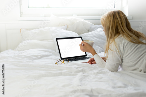 Blonde woman is sitting on the white bed and looks at the mockup laptop. Remote work. Covid 19. Young woman in on bed using laptop With blank mockup template on screen display. Work from home. White