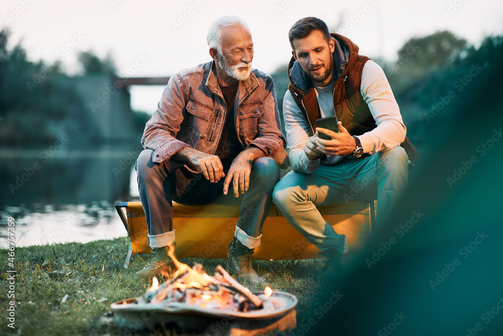 Mid adult man and his senior father use smart phone by campfire in nature. - obrazy, fototapety, plakaty 
