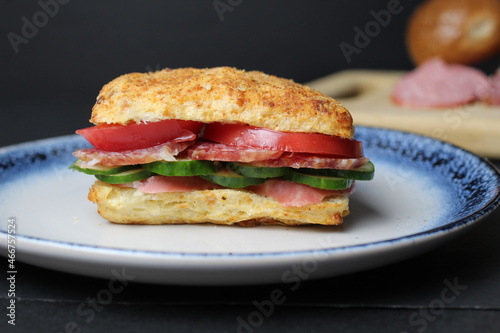 sandwich sandwich of white square bun with vegetables tomato cucumber sausage ham on a blue plate. in the background is a tray board with ingredients vegetables tomatoes on a black background. 