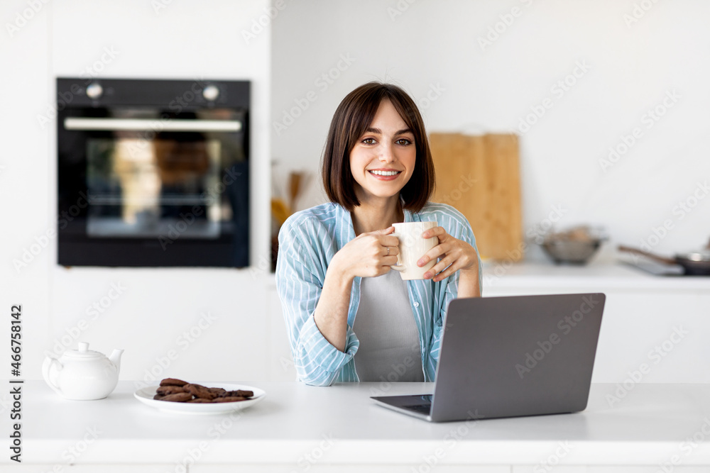 Portrait of happy lady using laptop and holding cup, drinking hot coffee in the morning, sitting in modern kitchen Portrait of happy lady using laptop and holding cup, drinking hot coffee in the morning, sitting in modern kitchen
