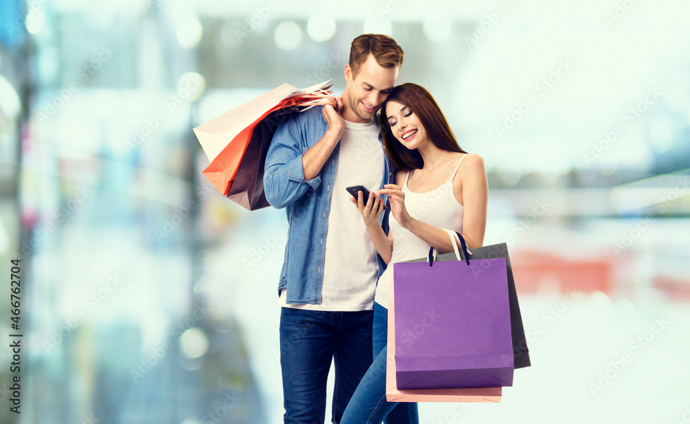 Love, holiday sales, shop, retail, consumer concept - happy couple with shopping bags, looking at mobile phone. Man and woman standing on blurred modern interior or mall background. Love, holiday sales, shop, retail, consumer concept - happy couple with shopping bags, looking at mobile phone. Man and woman standing on blurred modern interior or mall background.