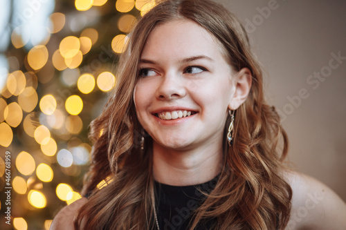 Portrait of an attractive young girl in a black evening dress against the background of a dressed up Christmas tree