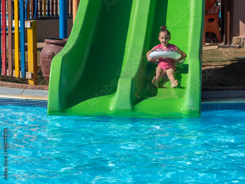 A pretty smiling little girl with a colorful inflatable ring goes down a green water slide in a pool with blue clear water in a water park. Summer vacation at the resort.