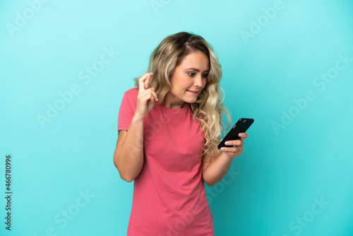 Young Brazilian woman isolated on blue background using mobile phone with fingers crossing