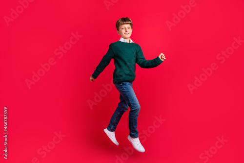 Photo of handsome sweet schoolboy dressed green sweater jumping high running smiling isolated red color background © deagreez