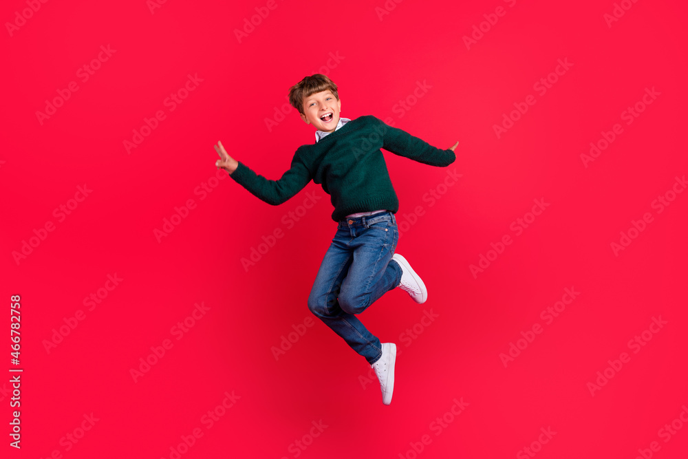 Photo of excited funny schoolboy dressed green sweater jumping high showing v-sign smiling isolated red color background