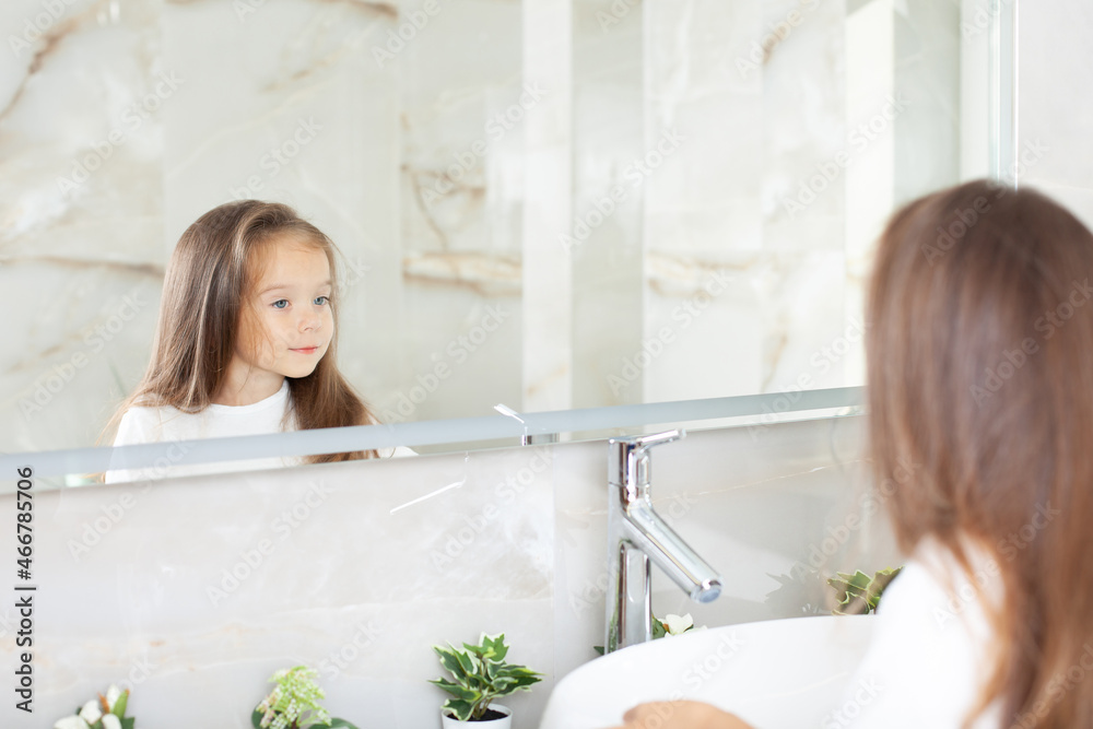 Portrait of a cute girl which looks in the mirror in a bright bathroom with a window. Hygiene. Morning routine. Purity. Like mother Portrait of a cute girl which looks in the mirror in a bright bathroom with a window. Hygiene. Morning routine. Purity. Like mother