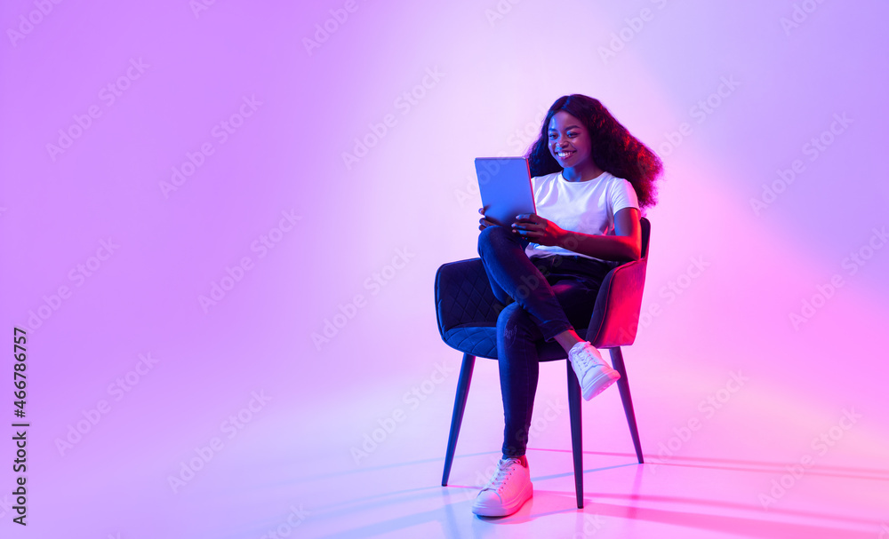 Cheerful black lady using tablet, studying or working remotely, sitting in armchair in neon light, copy space - obrazy, fototapety, plakaty 