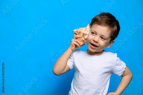 A boy listens to a shell on a blue background . Dreams of the sea. Article about vacation. Childhood. Baby and shell. Blue background.