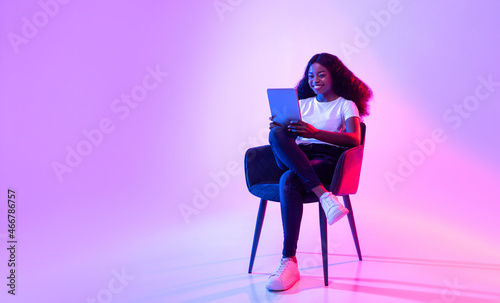 Cheerful black lady using tablet, studying or working remotely, sitting in armchair in neon light, copy space