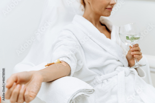Close up of calm female patient sitting with tube and needle during IV infusion. Girl holding glass with lemon water and relaxing photo