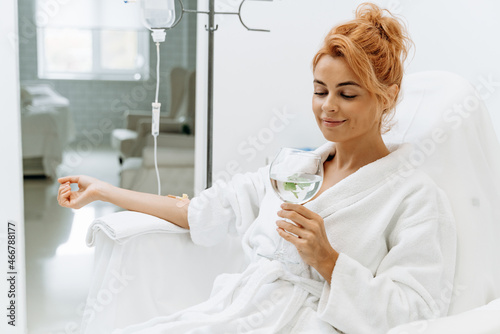 Waist up portrait view of the charming woman in white bathrobe sitting in armchair and receiving IV infusion. She is holding glass of lemon beverage and smiling photo