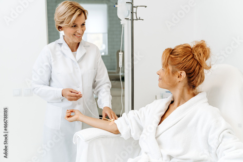 Side view portrait of attractive woman in white bathrobe sitting in armchair with pleasure smile while receiving IV infusion