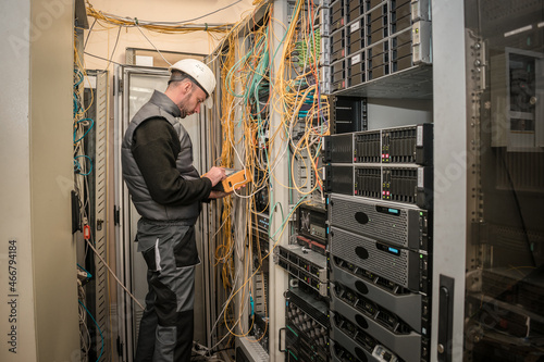 A technician works near racks with network equipment and lots of wires. An engineer in a white helmet is measuring the level of an optical signal in a server room