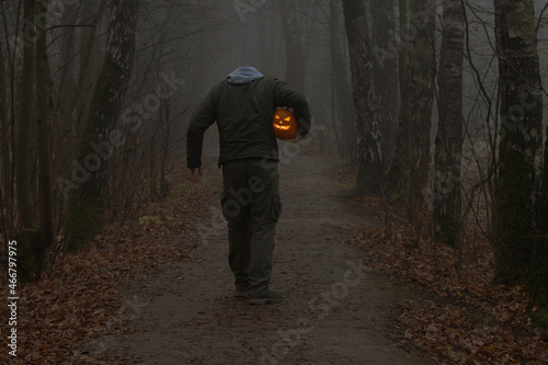 Headless man holds Jack O'Lantern Halloween pumpkin head under his arm and walks on countryside road in autumn foggy forest. Carved face on the pumpkin glows orange. Halloween costume theme.