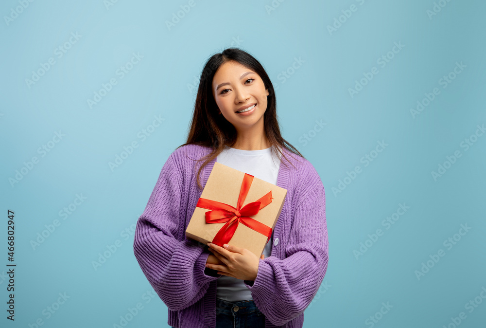 Young asian lady holding gift box and smiling to camera over blue studio background, free space