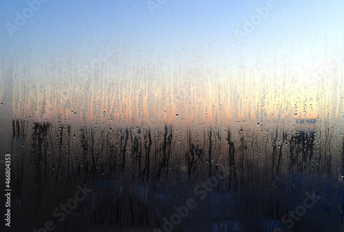 Sunrise. Sunset. Through a fogged window with dripping water drops, a clear blue sky and a red glow on the horizon are visible.