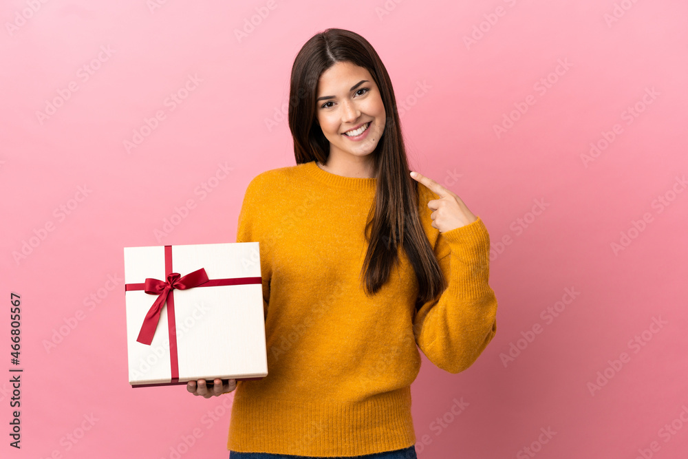 Teenager Brazilian girl holding a gift over isolated pink background giving a thumbs up gesture