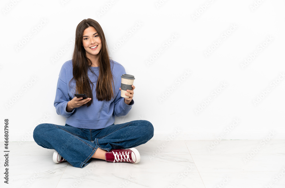 Young woman sitting on the floor holding coffee to take away and a mobile Young woman sitting on the floor holding coffee to take away and a mobile