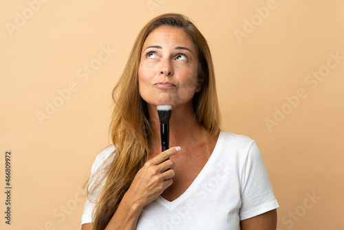 Middle age brazilian woman isolated on beige background holding makeup brush and thinking