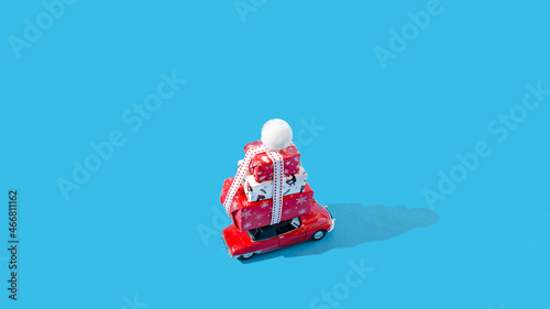 Fototapeta Naklejka Na Ścianę i Meble -  A red car with a pile of wrapped Christmas presents on the roof. Pastel blue background. Christmas or New Year celebration gifts creative concept. Card or banner or invitation design.